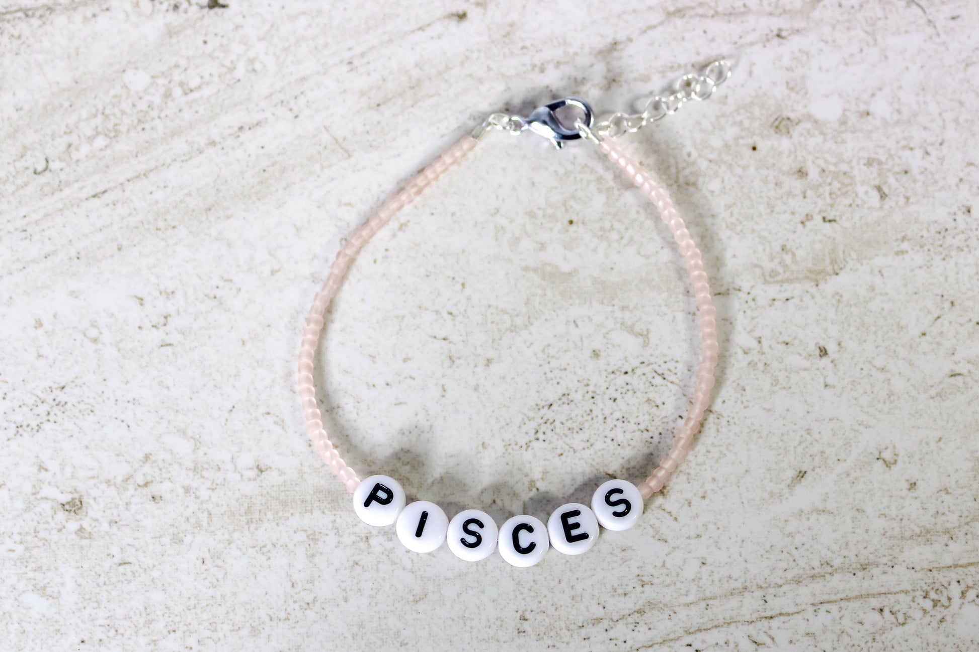 A pale pink colored seed bead bracelet with Pisces in round letter beads and silver lobster clasp and extender chain.