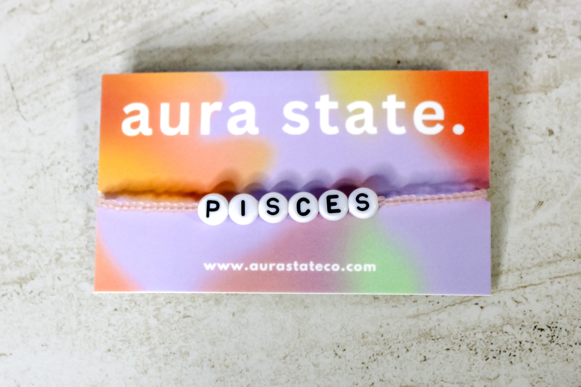 A pale pink beaded bracelet with plastic round beads that spell Pisces on a rectangular card that says aura state.