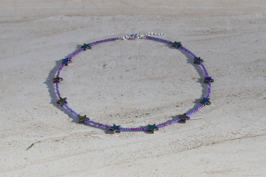 A necklace made out of purple seed beads with rainbow metal stars spaced evenly throughout.  Features a silver lobster claw clasp and chain extender.