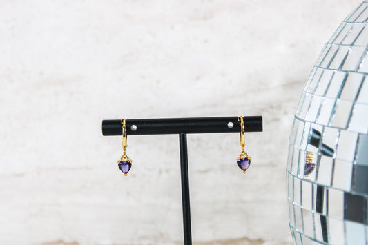 A pair of gold huggie hoop earrings with small purple cubic zirconia heart charms are displayed on a t bar.