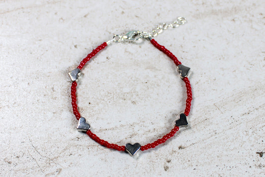 A bracelet made out of red seed beads with silver metal hearts spaced evenly throughout.  Features a silver lobster claw clasp and chain extender.