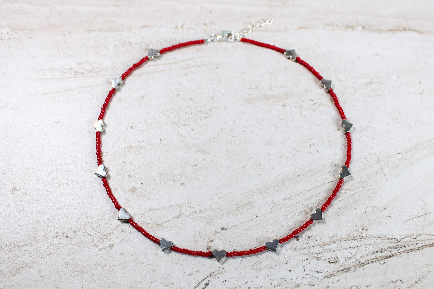 A necklace made out of red seed beads with silver metal hearts spaced evenly throughout.  Features a silver lobster claw clasp and chain extender.