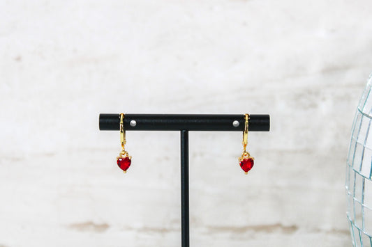 A pair of gold huggie hoop earrings with small red cubic zirconia heart charms are displayed on a t bar.