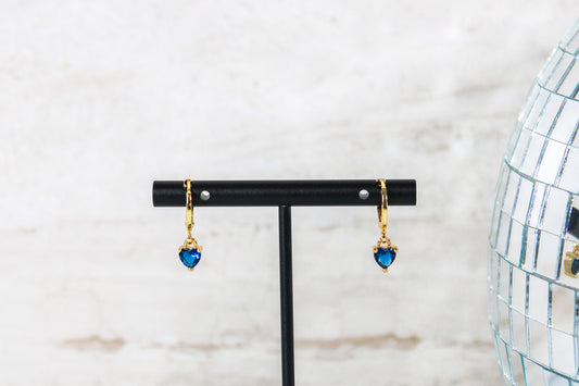 A pair of gold huggie hoop earrings with small blue cubic zirconia heart charms are displayed on a t bar.