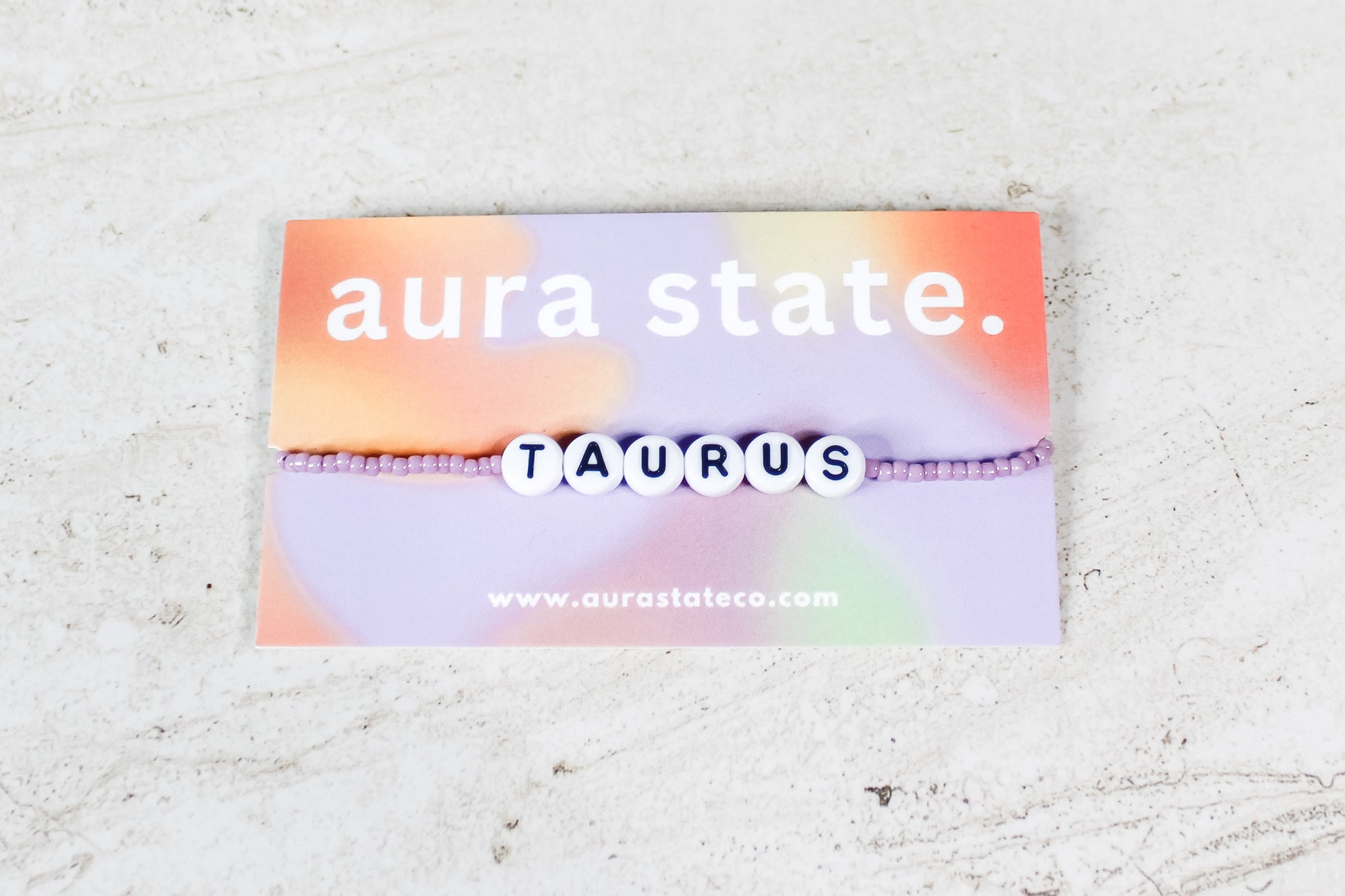 A shiny mauve beaded bracelet with plastic round beads that spell Taurus on a rectangular card that says aura state.