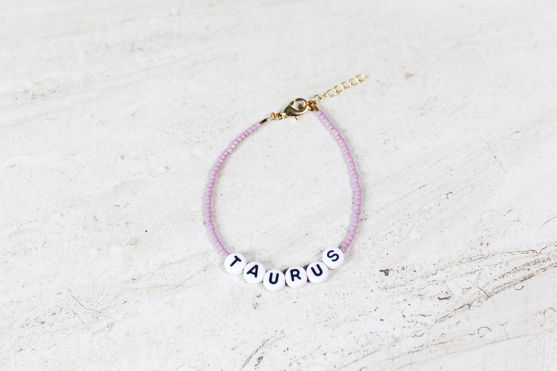 A shiny mauve colored seed bead bracelet with Taurus in round letter beads and gold lobster clasp and extender chain.