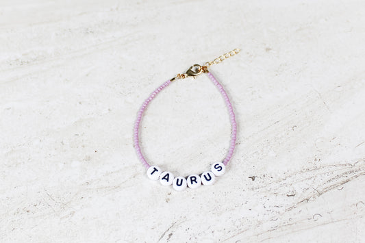 A shiny mauve colored seed bead bracelet with Taurus in round letter beads and gold lobster clasp and extender chain.