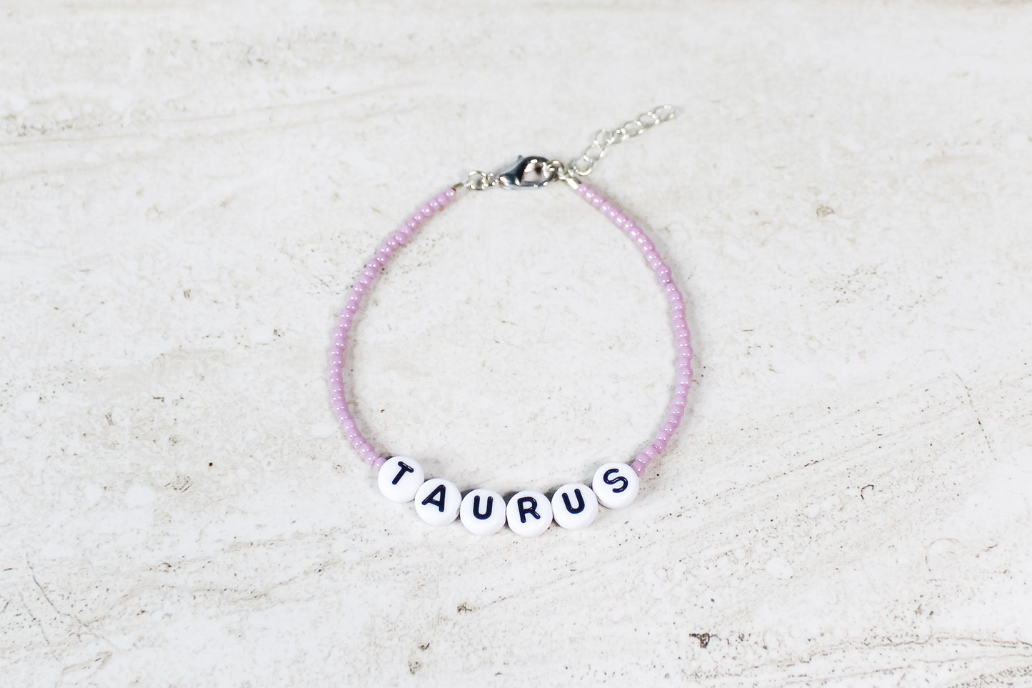 A shiny mauve colored seed bead bracelet with Taurus in round letter beads and silver lobster clasp and extender chain.