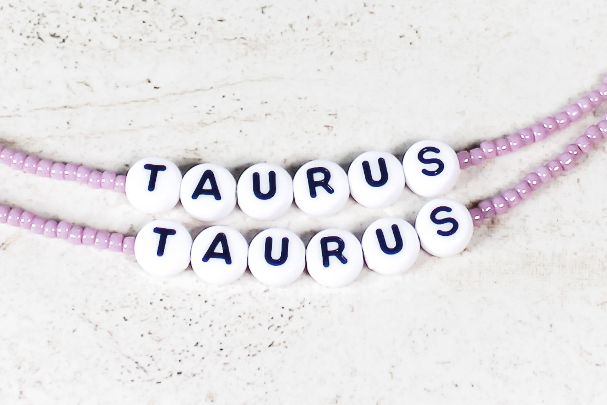 Two bracelets made out of shiny mauve seed beads with plastic beads that spell out Taurus.