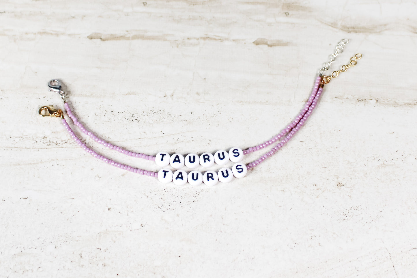 Two bracelets made out of shiny mauve seed beads with plastic beads that spell out Taurus.