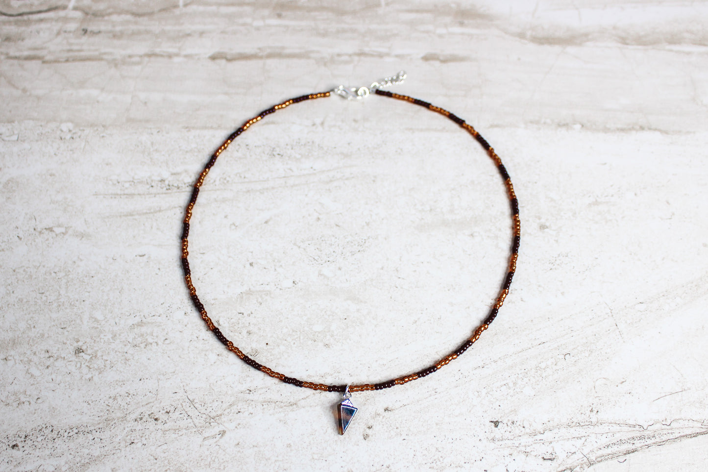 A handmade necklace with brown and copper seed beads, finished with a silver extender chain and a triangle-shaped tortoiseshell charm at the center.