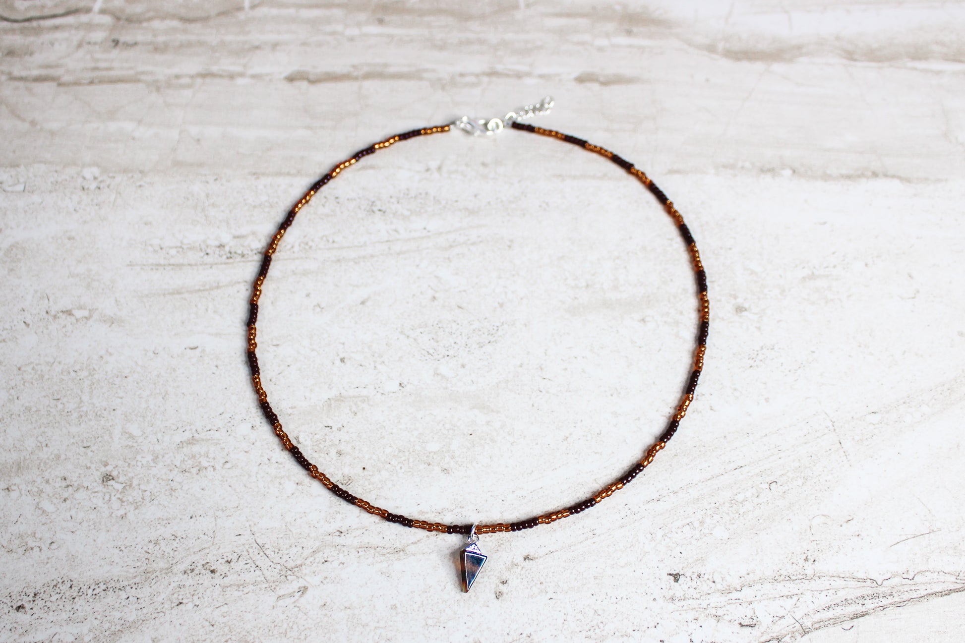 A handmade necklace with brown and copper seed beads, finished with a silver extender chain and a triangle-shaped tortoiseshell charm at the center.