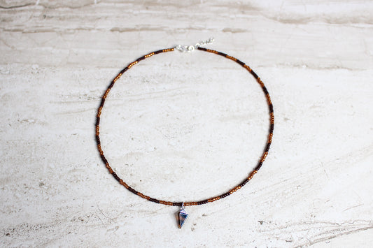 A handmade necklace with brown and copper seed beads, finished with a silver extender chain and a triangle-shaped tortoiseshell charm at the center.