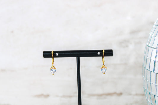 A pair of gold huggie hoop earrings with small white cubic zirconia heart charms are displayed on a t bar.